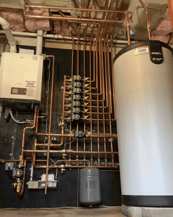 Mechanical room with a water heater, boiler system, and numerous copper pipes and valves neatly arranged on the wall. Insulation is visible on the ceiling.