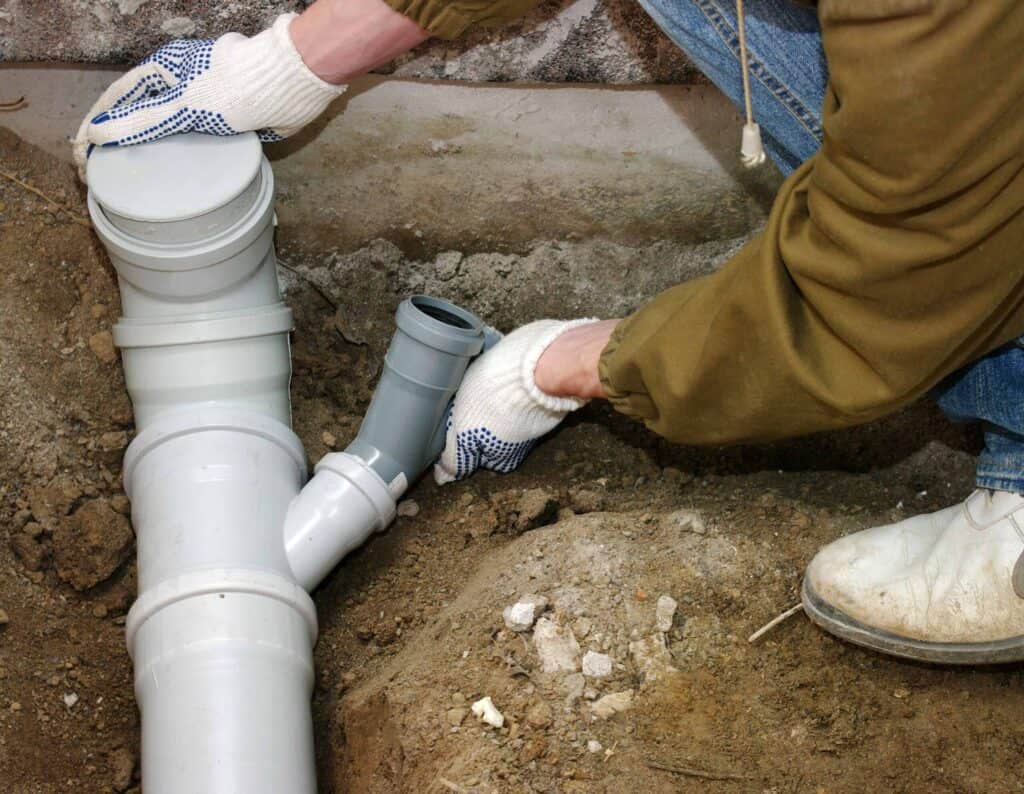 Person in gloves working on PVC plumbing pipes buried in dirt.