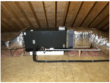 HVAC unit installed in an attic, surrounded by insulated air ducts and wooden flooring, with sloped wooden ceiling above.