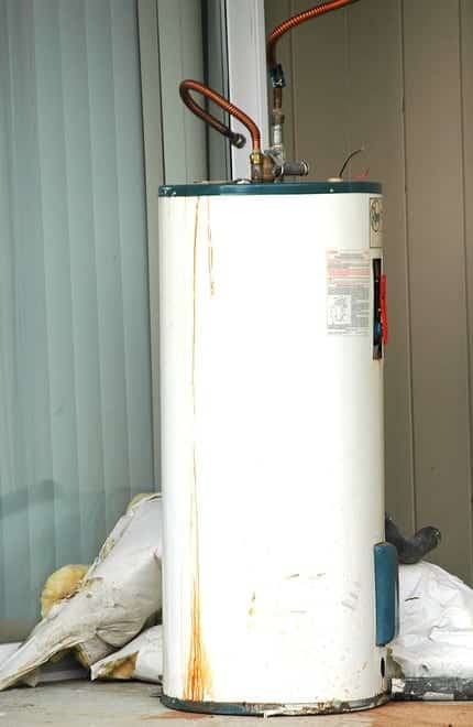 A white, cylindrical water heater stands upright outdoors, with visible rust stains and pipes connected at the top.