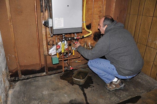 A person in a gray hoodie examines pipes connected to a wall-mounted boiler in a basement. Water is on the floor, and a small tray is placed underneath the pipes.