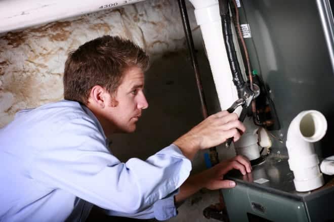 Person in a blue shirt using a wrench to repair HVAC equipment in a basement.