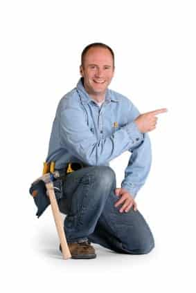 A person in a blue shirt and jeans kneels, smiling and pointing to the right. A hammer and tools are in their tool belt.