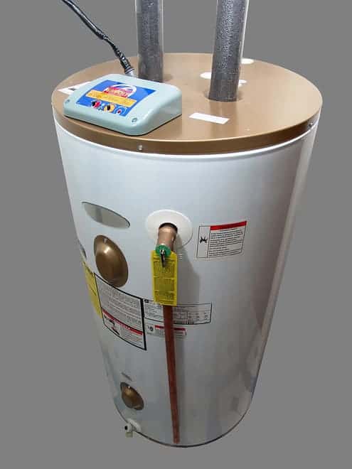 A white cylindrical water heater with a beige top, featuring various labels, a control box on top, two silver pipes, and a protruding copper pipe with a green tag.