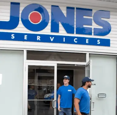 Two employees in blue uniforms exit a building with large "Jones Services" signage above the entrance, expertly carrying tools for furnace installation.