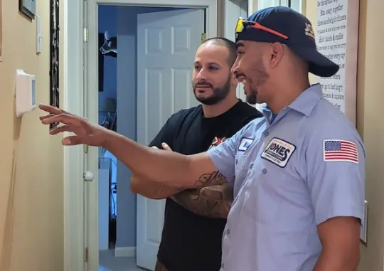 Two men stand in a hallway. One, in a uniform and cap, discusses general heating repair as he points to a thermostat on the wall, while the other man listens attentively.