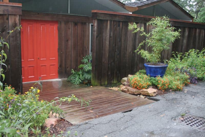 Rustic backyard garden with wooden pathway, vibrant potted plants, and natural stone accents, designed by John Russell Landscape Architect for aesthetic and functional outdoor space enhancement.
