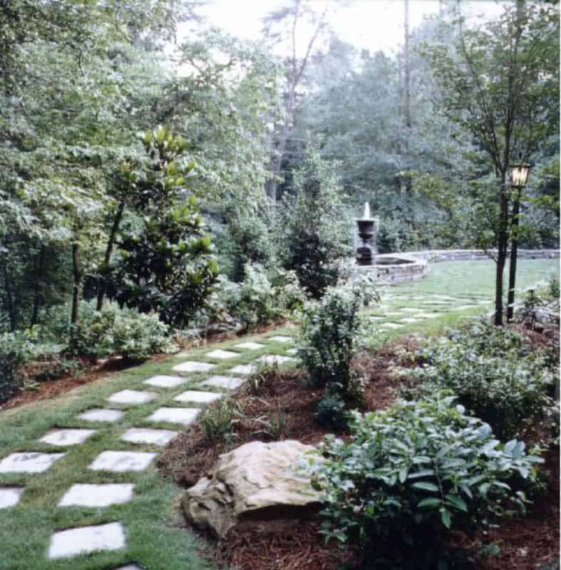 Stone pathway through lush garden with mature trees and shrubs, outdoor lighting, and water feature, designed by John Russell Landscape Architect, emphasizing natural beauty and landscape harmony.