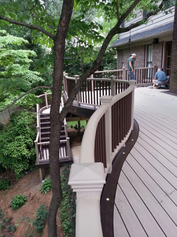 Elegant outdoor wooden deck with curved railing and staircase, surrounded by lush greenery, designed by John Russell Landscape Architect, perfect for outdoor living and entertaining in a natural setting.