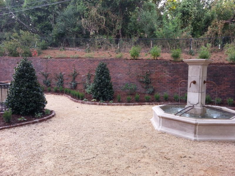 Aesthetic backyard landscape with a decorative stone fountain, well-maintained shrubs, and a brick retaining wall, designed by John Russell Landscape Architect.