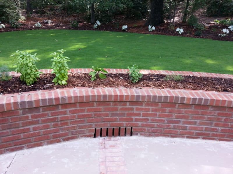 Lush garden bed with healthy basil and other plants bordered by classic red brick retaining wall, complemented by a well-maintained green lawn and garden in the background.