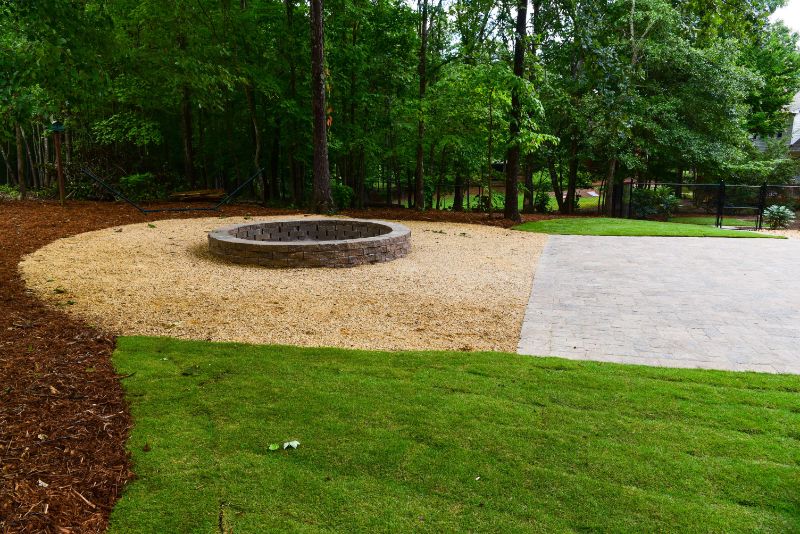 Round stone fire pit with gravel seating area in landscaped backyard with lush trees and grass, designed by John Russell Landscape Architect.