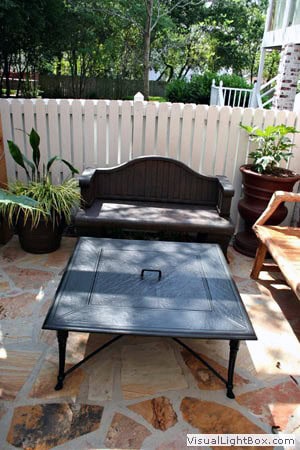 Aesthetic outdoor seating area with black metal table, potted plants, and wooden bench, surrounded by white picket fence and lush greenery, designed by John Russell Landscape Architect.