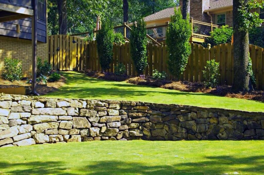 Stone retaining wall with landscaped yard and wooden fence in a backyard garden, outdoor landscape design by John Russell Landscape Architect, lush greenery, modern xeriscape irrigation, residential outdoor space.