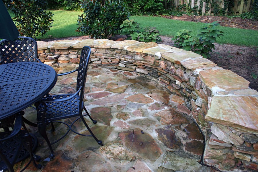 A beautiful stone outdoor patio with a curved stone planter and wrought iron furniture, designed by John Russell Landscape Architect. Perfect for relaxing and entertaining in a lush garden setting.
