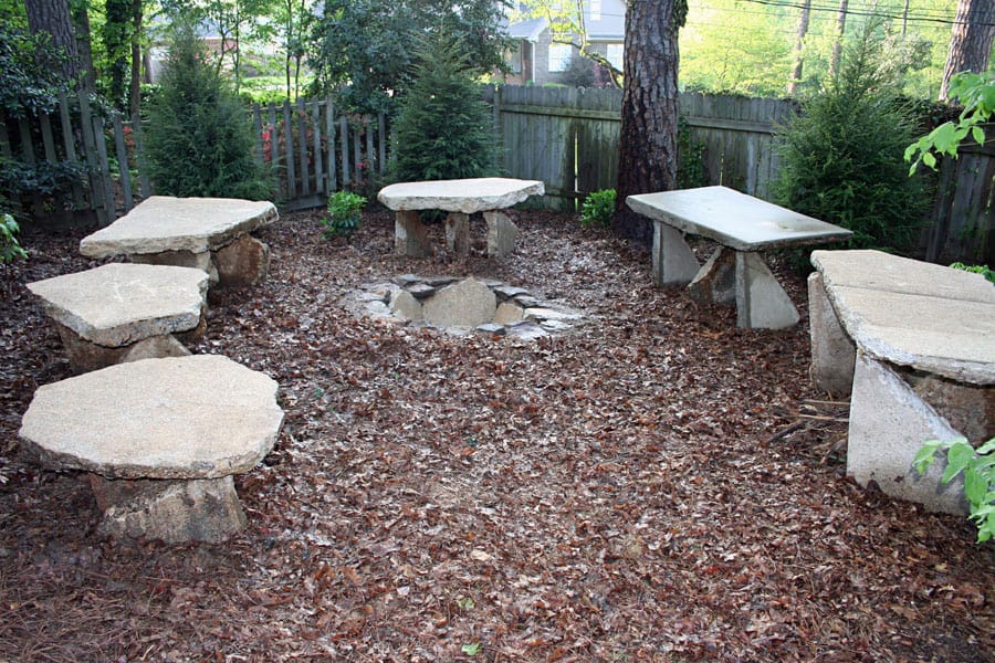 Stone fire pit circle with rustic stone benches in landscaped backyard garden, ideal for outdoor gatherings and relaxation, created by John Russell Landscape Architect.