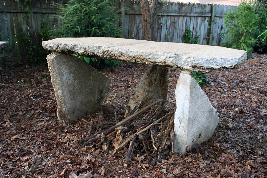 Natural stone outdoor table with rustic design, featuring a large flat stone top supported by irregular stone legs, surrounded by garden foliage.