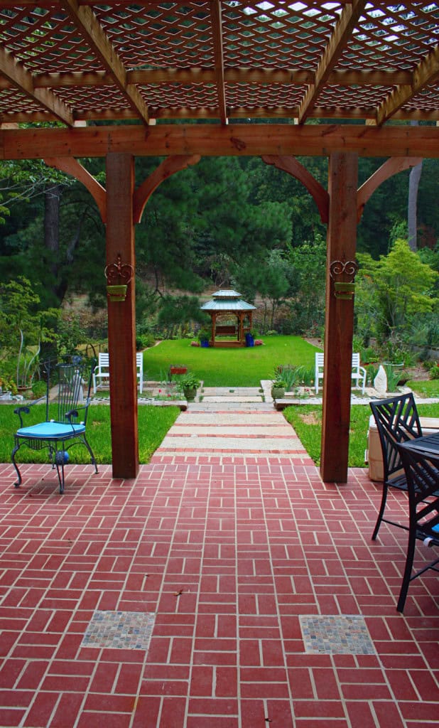 Elegant wooden pergola with lattice roof over red brick patio leading to lush green backyard with a gazebo, designed by John Russell Landscape Architect.