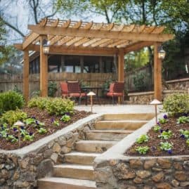 Natural stone landscape stairs with lush flower beds leading up to a wooden pergola patio area in a landscaped backyard.