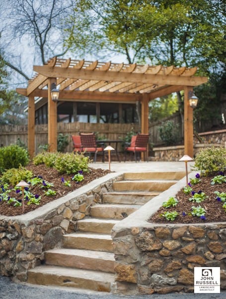 Natural stone landscape stairs with lush flower beds leading up to a wooden pergola patio area in a landscaped backyard.