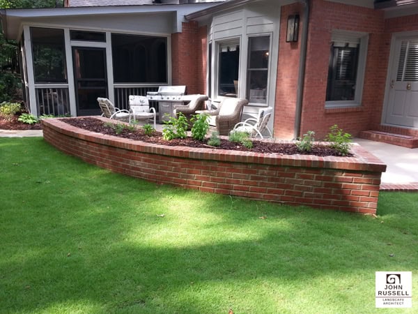 Brick retaining wall landscape design with outdoor dining area and lush green lawn, enhancing backyard curb appeal and functional outdoor living space by John Russell Landscape Architect.