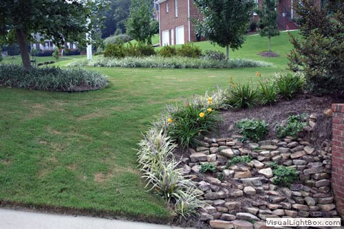 Lush landscaped yard with stone retaining wall, vibrant plants, and well-manicured grass, designed by John Russell Landscape Architect in a suburban setting.