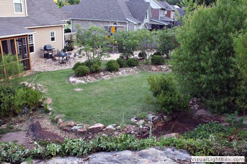 Lush backyard landscaping with vibrant plants, mature trees, and stone features designed by John Russell Landscape Architect in a suburban setting.
