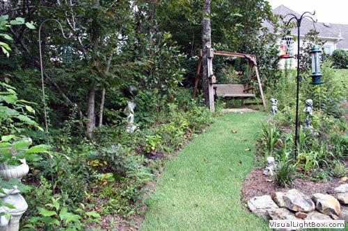 Lush garden landscape design with a stone pathway, decorative statues, and wooden swing set created by John Russell Landscape Architect.