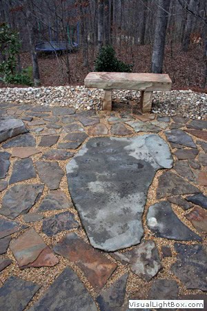Handcrafted natural stone patio with flagstone pathway and earthy landscape design in a wooded backyard setting.