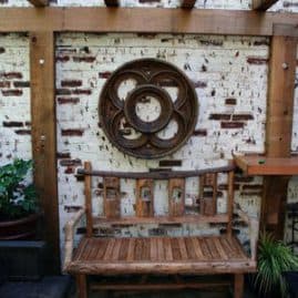 Modern outdoor patio with rustic brick wall, wooden garden bench, and contemporary wall sculpture, showcasing landscape architecture and outdoor living design by John Russell Landscape Architect.