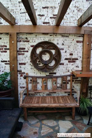 Modern outdoor patio with rustic brick wall, wooden garden bench, and contemporary wall sculpture, showcasing landscape architecture and outdoor living design by John Russell Landscape Architect.