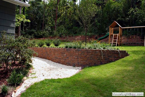 Lush backyard landscape featuring a brick retaining wall, gravel pathway, and a wooden playset amid mature trees and greenery, designed by John Russell Landscape Architect.
