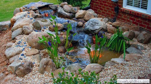 Natural stonewater feature with cascading waterfall, lush greenery, and blooming plants, designed by John Russell Landscape Architect for outdoor garden aesthetics.
