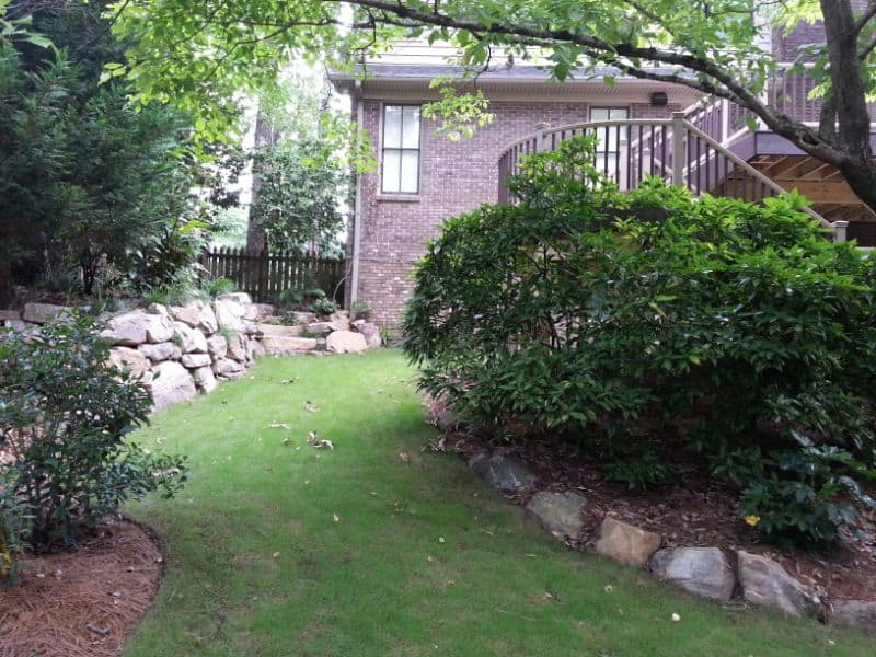 Lush backyard landscape design featuring green shrubs, stone borders, and a well-maintained lawn with a brick house in the background, created by John Russell Landscape Architect.
