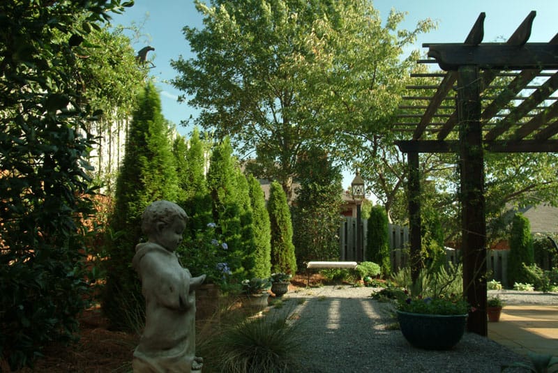 Lush backyard landscape design featuring mature trees, decorative sculptures, and a wooden pergola, created by John Russell Landscape Architect, showcasing expert outdoor space planning and lush greenery.