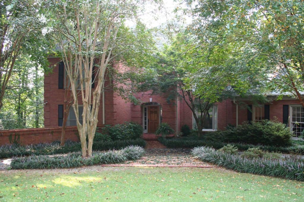 Lush landscaped front yard with mature trees and ornamental plants surrounding a historic brick house, showcasing expert landscape architecture.