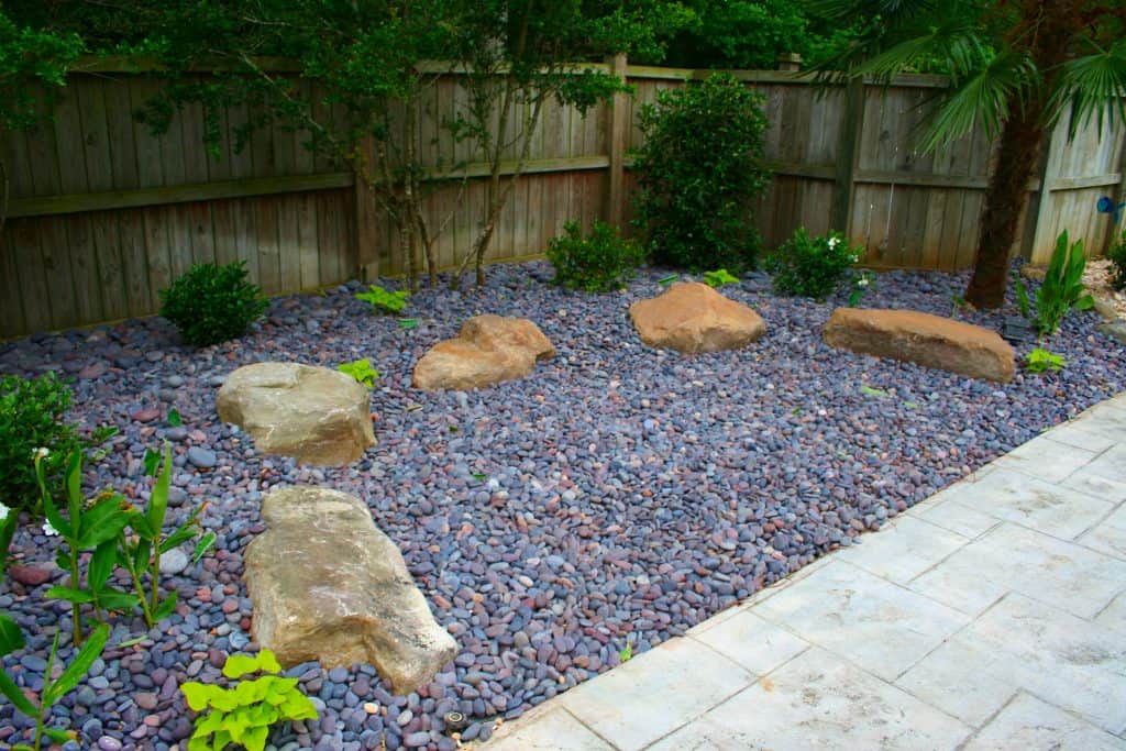 Large decorative landscape stones in a minimalist backyard design with purple gravel and lush greenery, showcasing professional landscape architecture by John Russell Landscape Architect.