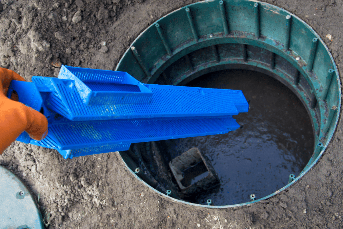 A person wearing an orange glove holds a blue plastic filter above an open circular manhole filled with murky water, surrounded by soil—suggesting septic tank maintenance is underway.
