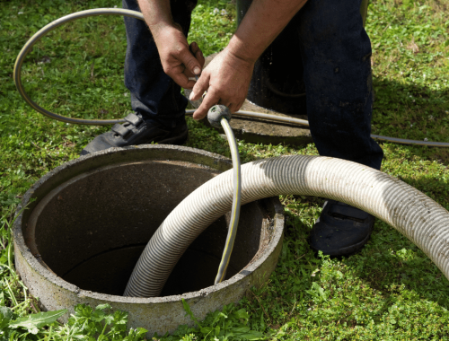 A person is holding hoses near the opening of an outdoor septic tank, preparing for septic system service or cleaning. The surrounding area is grassy.
