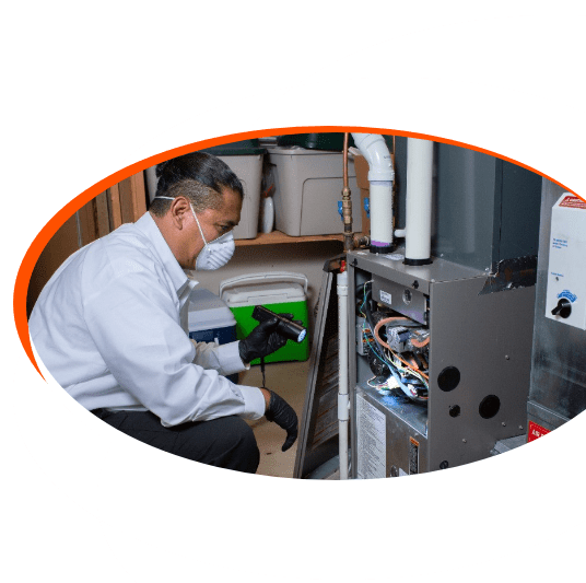 A technician wearing a mask and gloves inspects the inside of a furnace unit with a flashlight in a utility room.
