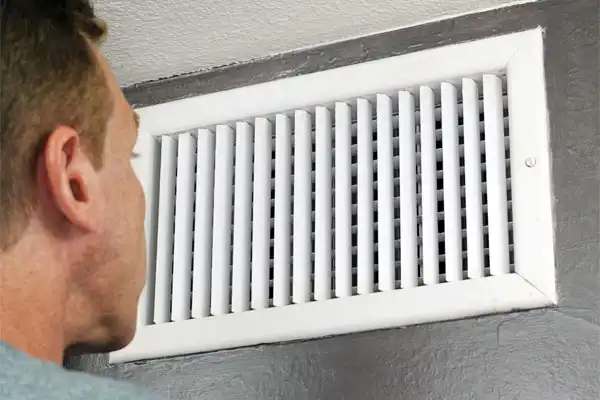 A person in Kamloops, BC, closely inspecting a white air vent mounted on a gray wall, possibly checking for areas needing air duct sealing.