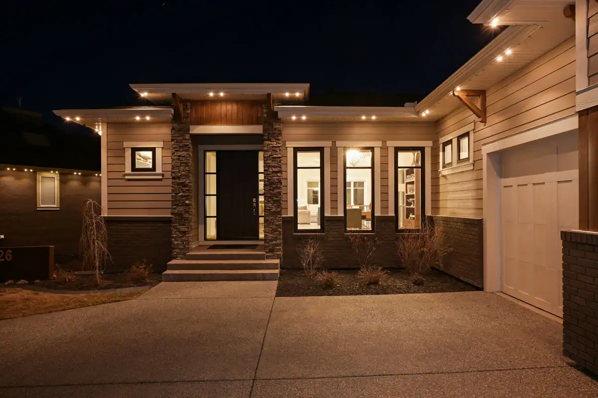 Modern house exterior at night with stone accents, large windows, a lit entrance, and an attached garage. The driveway and front landscaping are visible, reflecting the contemporary style you'll discover in our about us section.