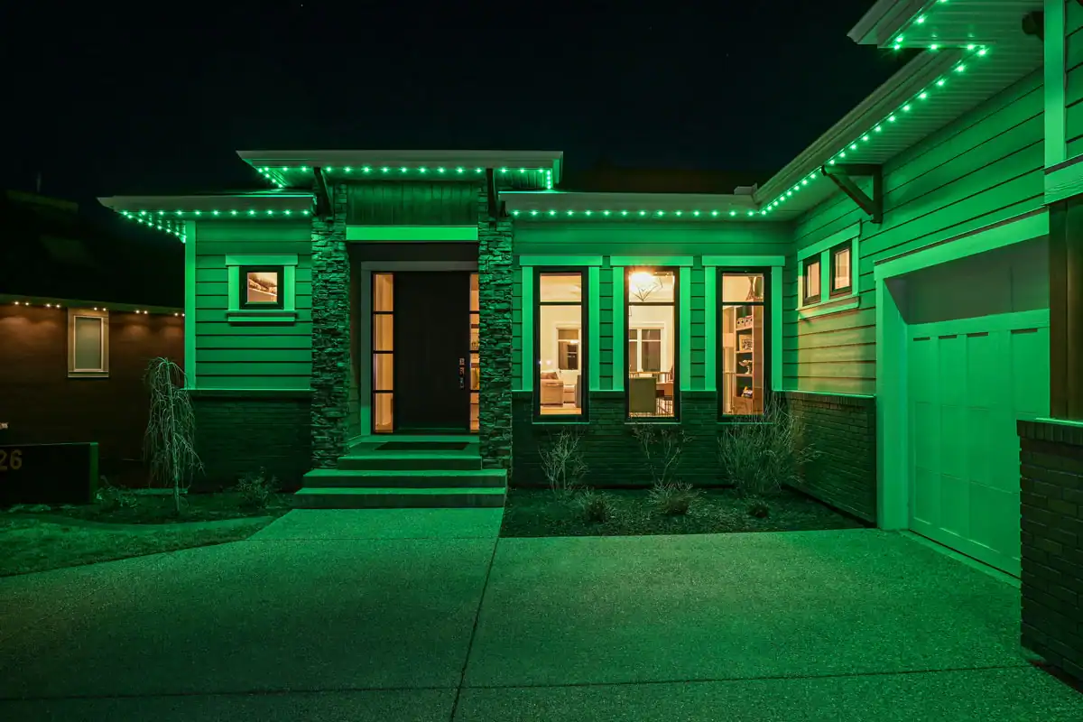 Modern house exterior at night illuminated by green LED lights, with large windows showing a well-lit interior and a garage on the right—learn more about us through our innovative home designs.