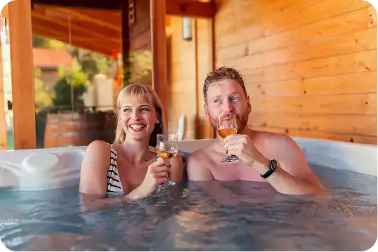 Two people sit in a hot tub, smiling and holding glasses of wine, with a wooden cabin structure in the background.