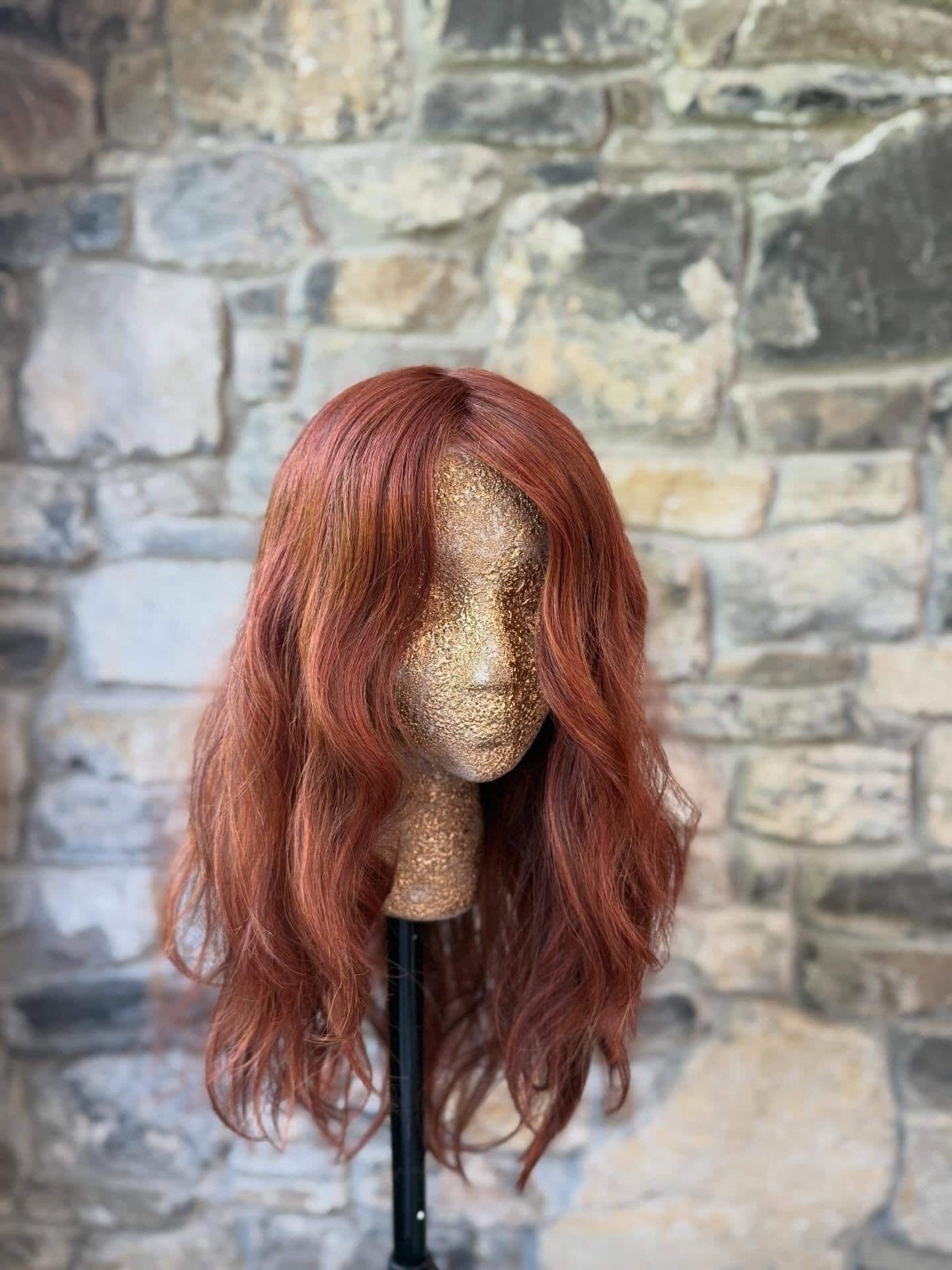 Red wavy hair wig on a mannequin against a stone wall background.