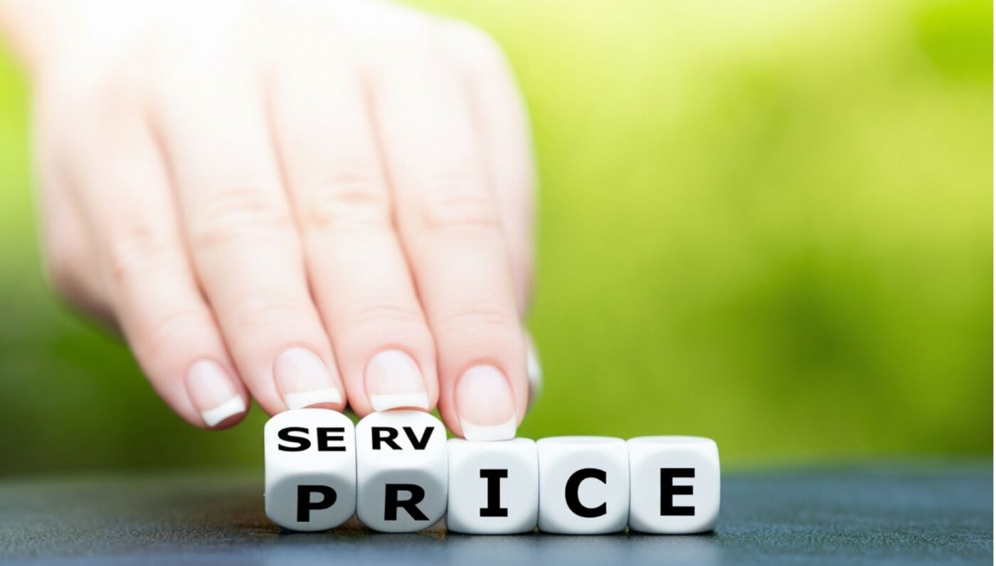 Hand berührt kleine Würfel mit dem Wort „Price“ auf einem Tisch, vor unscharfem grünem Hintergrund.