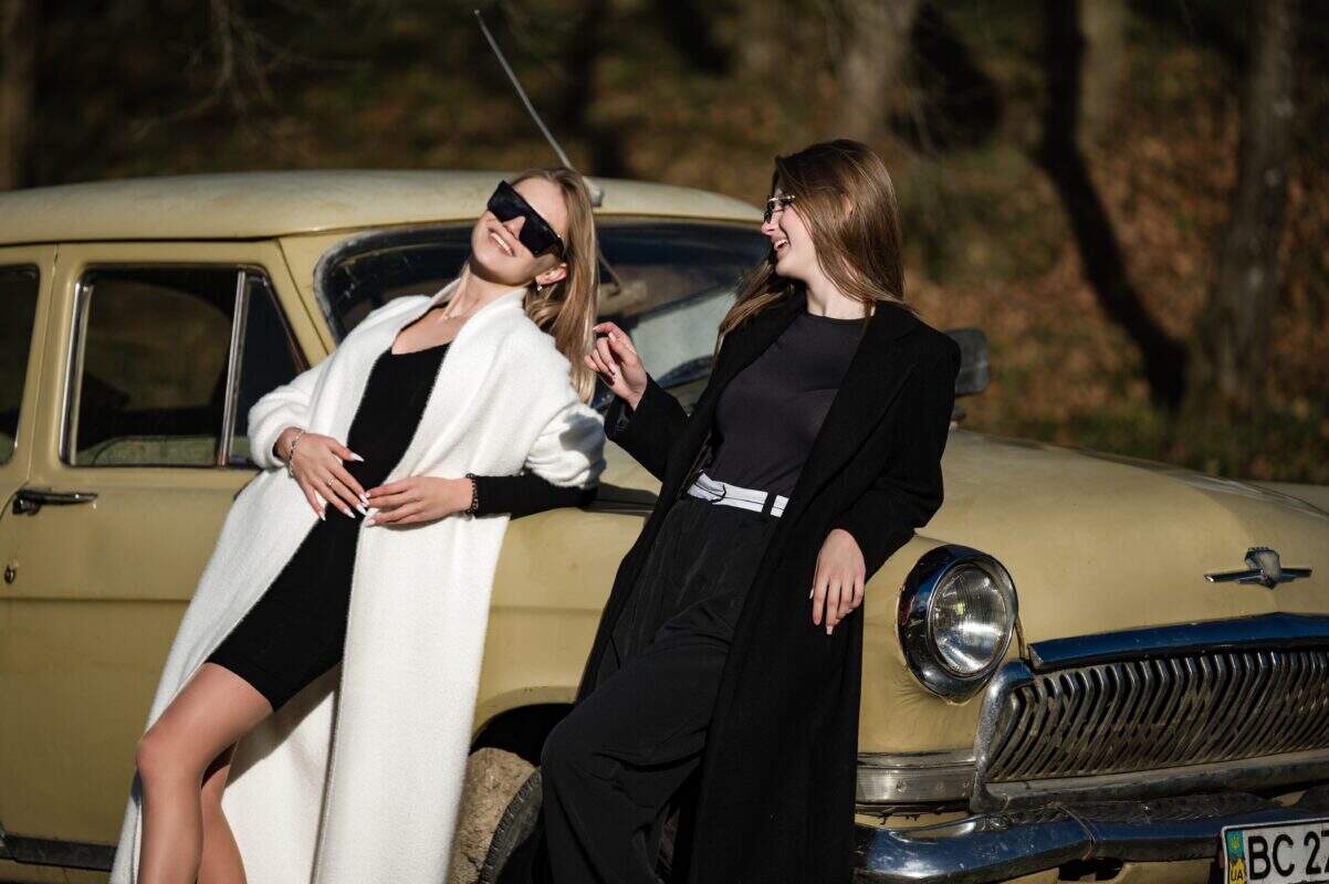 Zwei Frauen posieren für ein Selfie neben einem klassischen Auto in einer stilvollen Outdoor-Szene.