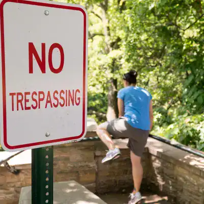 No trespassing sign blocking access to private property with a person climbing over a fence in the background.
