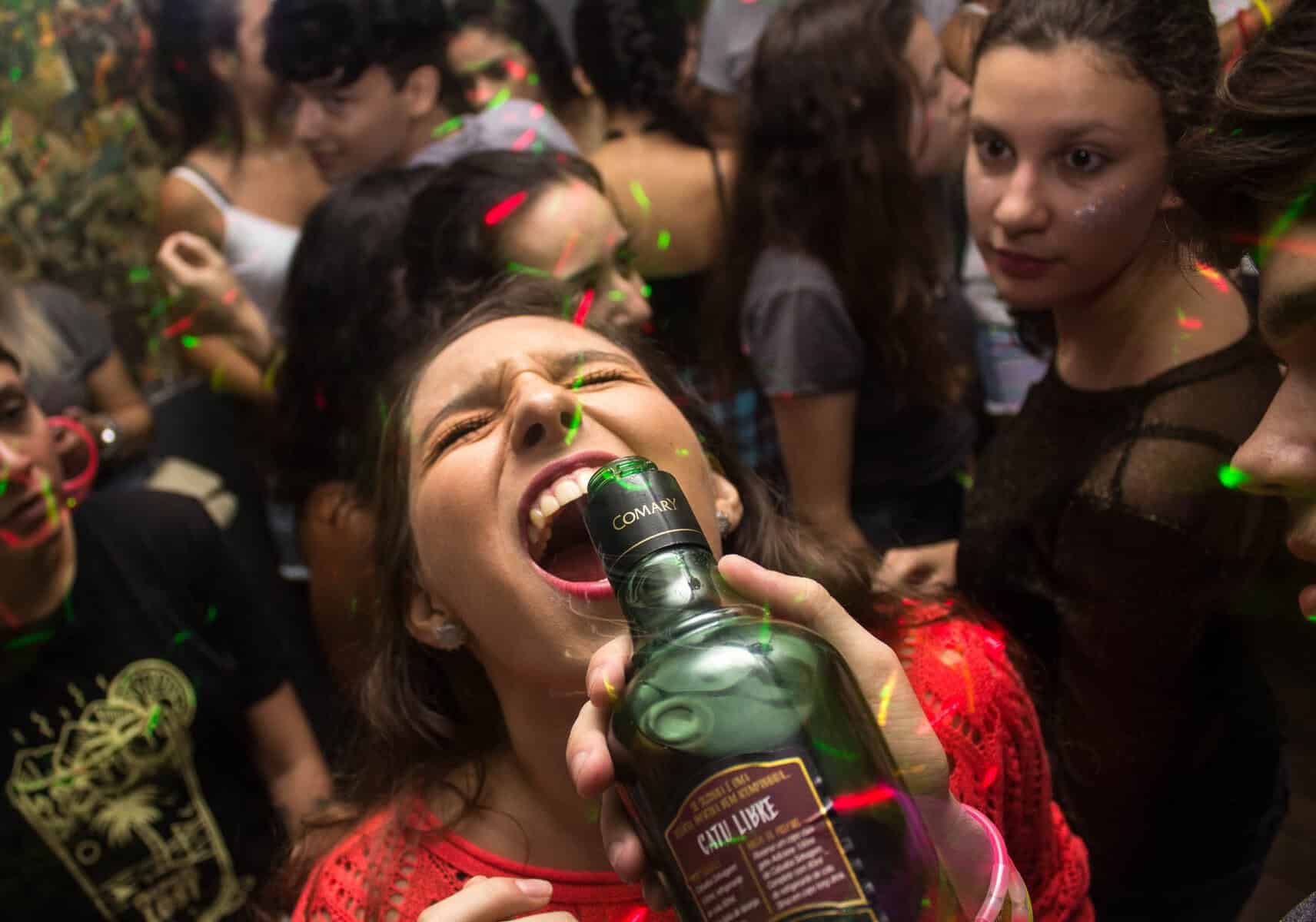 A woman partying enthusiastically surrounded by friends at a lively celebration, holding a bottle of liquor with colorful party lights and confetti in the background.