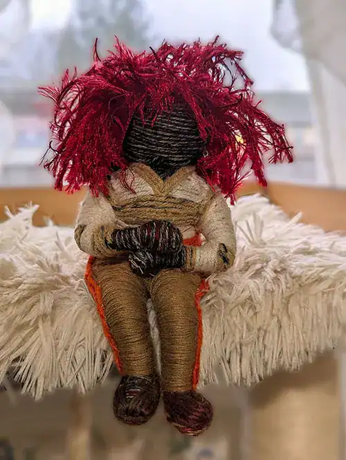 A voodoo art doll with red hair who wears a tracksuit with one orange stripe down each leg and a V-neck top with long sleeves